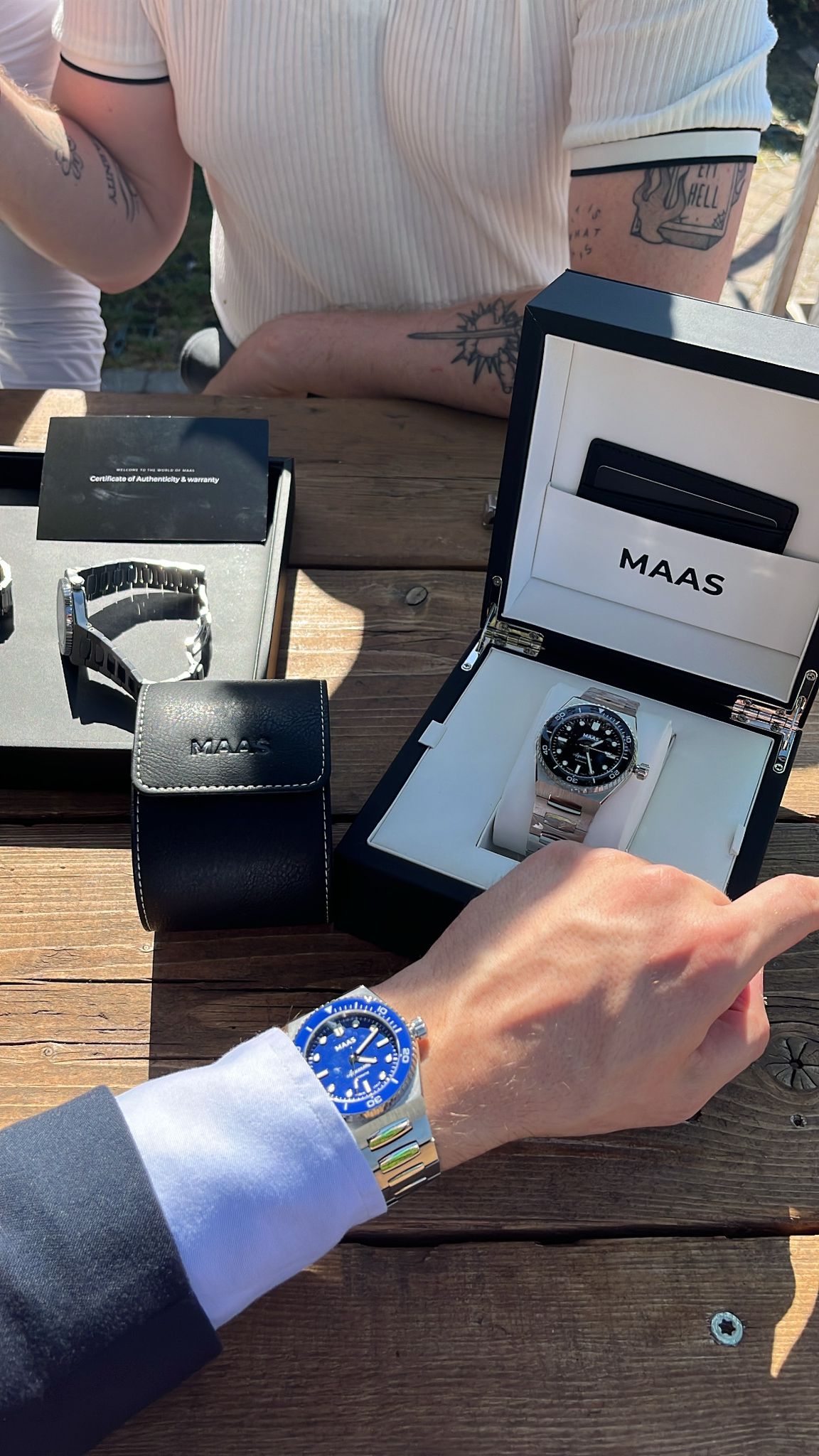 Person holding a blue watch with another watch in a 'MAAS' branded box on a wooden surface.
