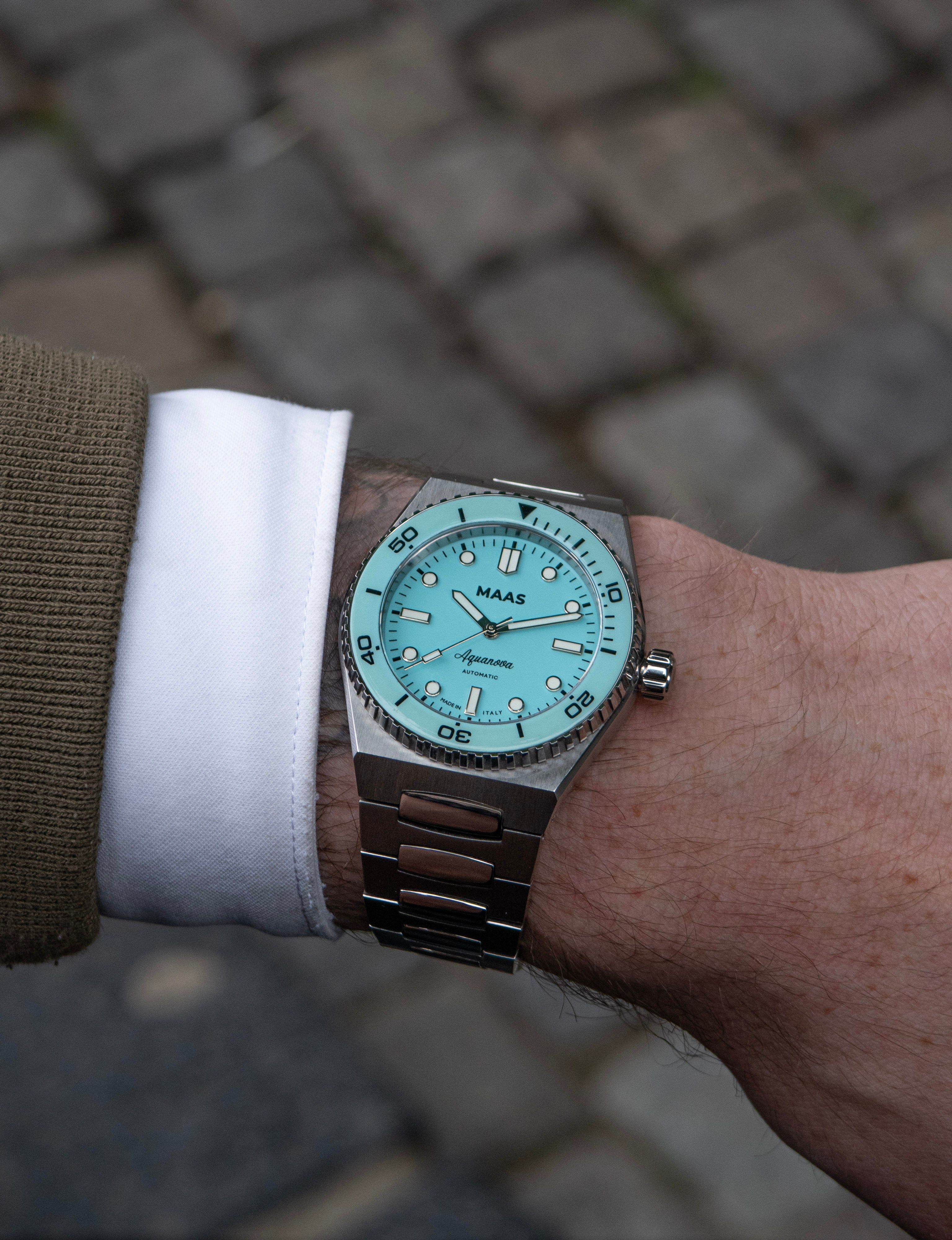 Close-up of a wrist wearing a watch with a blue face and brown strap on a blurred pavement background.
